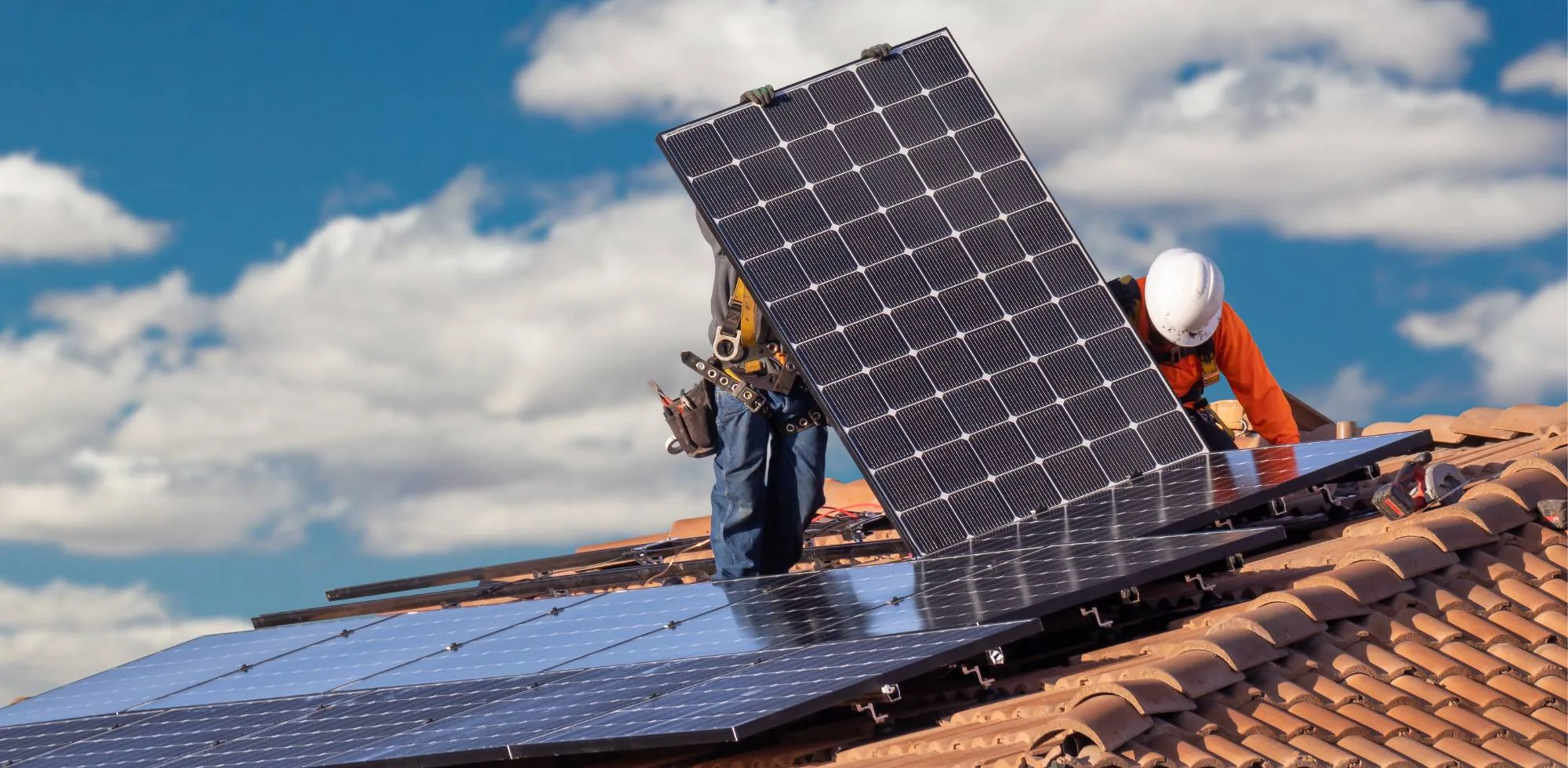 Two installers in harnesses lifting a monocrystalline module into position on a tiled pitched roof, mounting rails already fixed beneath it
